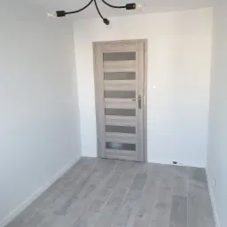 Wnętrze po remoncie w Rudzie Śląskiej: szare panele podłogowe imitujące drewno, jasnoszare drzwi z przeszkleniami i nowoczesna czarna lampa sufitowa z odsłoniętymi żarówkami na tle białych ścian.