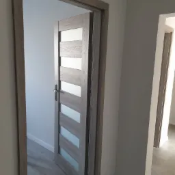 Otwarta, wewnętrzna, drewniana drzwi z przeszkleniami w odcieniach szarości, widok z perspektywy przechodzącego przez próg, z widocznym fragmentem podłogi i kolejnymi drzwiami w głębi.