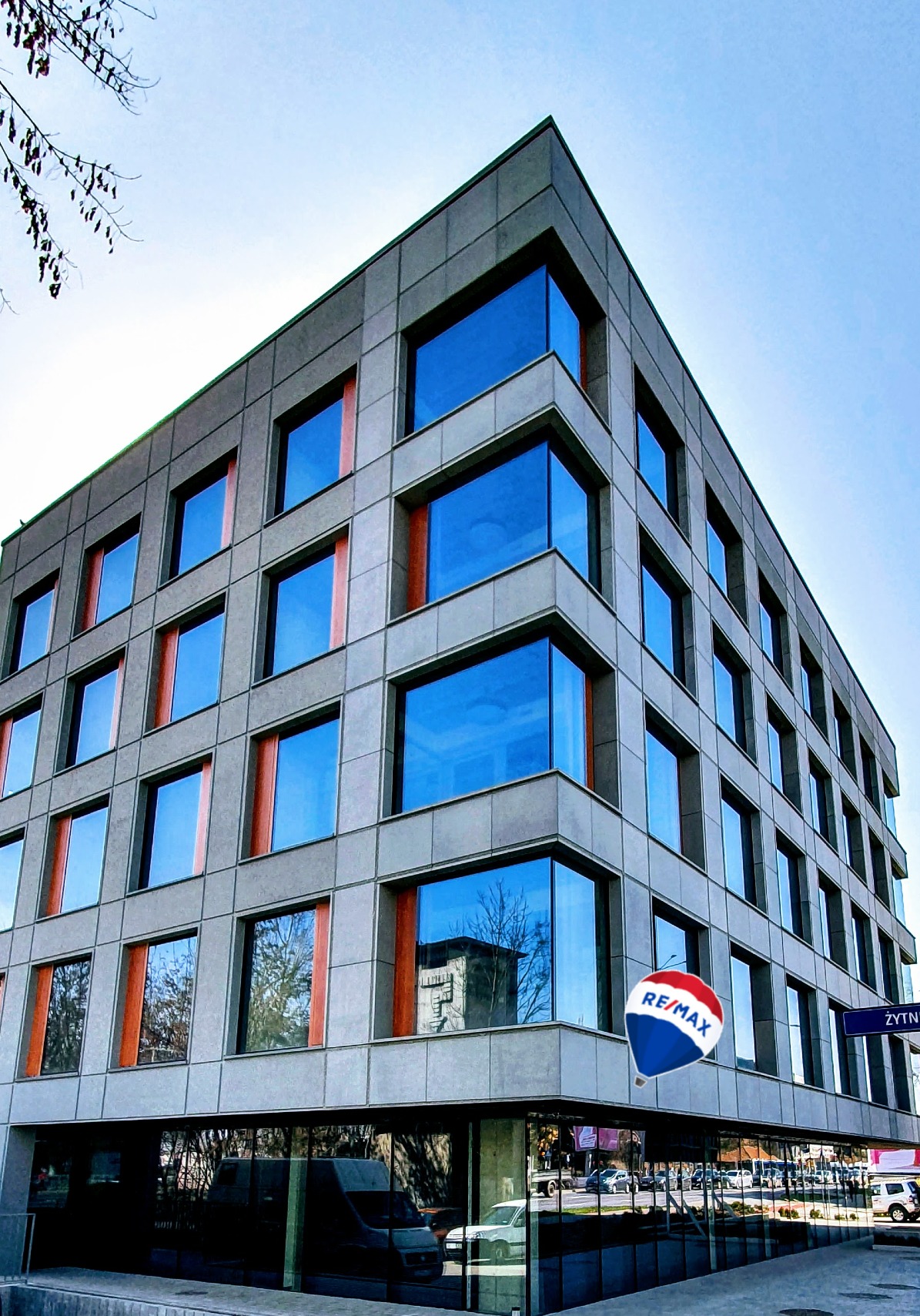 Nowoczesny budynek biurowy z szarych paneli z licznymi oknami odbijającymi niebo i drzewa, z logo RE/MAX w formie balonu w rogu.