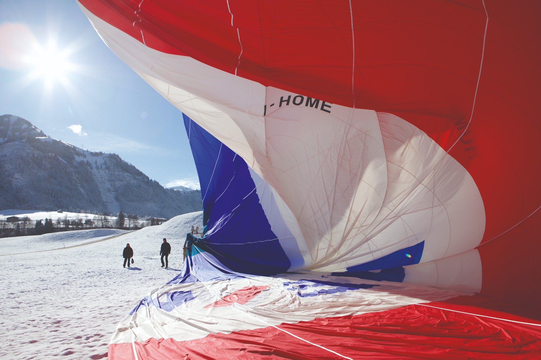 Wnętrze balonu na gorące powietrze z napisem '- HOME' na tle zasnieżonego krajobrazu górskiego i spacerujących osób.