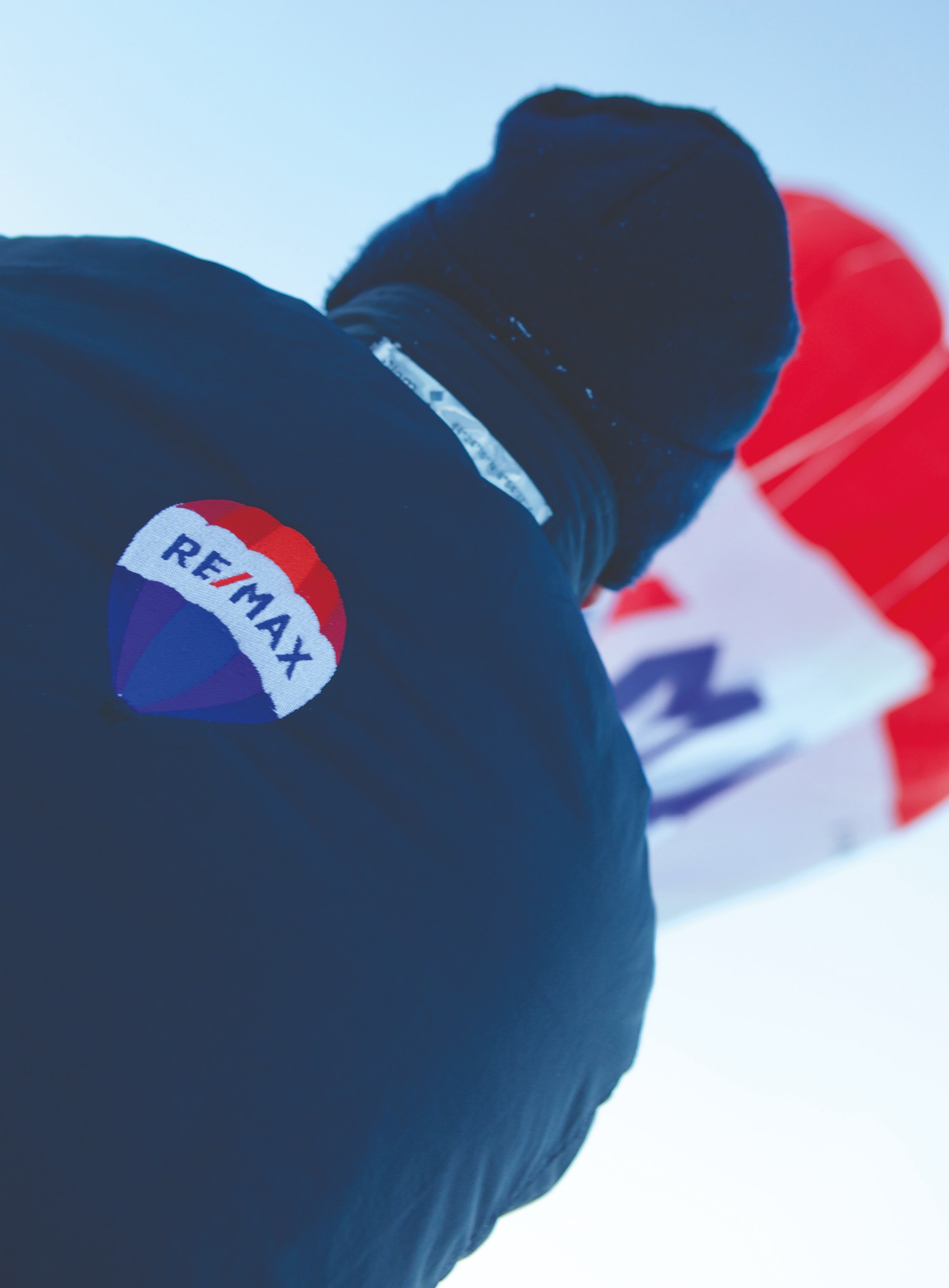 Osoba w ciemnej kurtce i czapce z naszywką RE/MAX, w tle częściowo widoczny czerwono-biały obiekt przypominający balon.