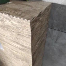 Wykończona obudowa WC w łazience z płytkami imitującymi drewno, widoczne narzędzia glazurnicze i fragment szarej ściany z płytek.