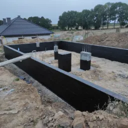 Przygotowany, zaizolowany fundament pod budowę domu jednorodzinnego, widoczne zbrojenia słupów, w tle częściowo wybudowany budynek i drzewa.