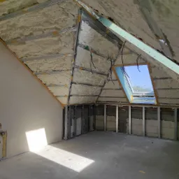 Poddasze w stanie surowym otwartym, ocieplone pianą natryskową, z widoczną konstrukcją ścian działowych z profili metalowych i oknem dachowym, wpadające promienie słoneczne na betonową posadzkę.