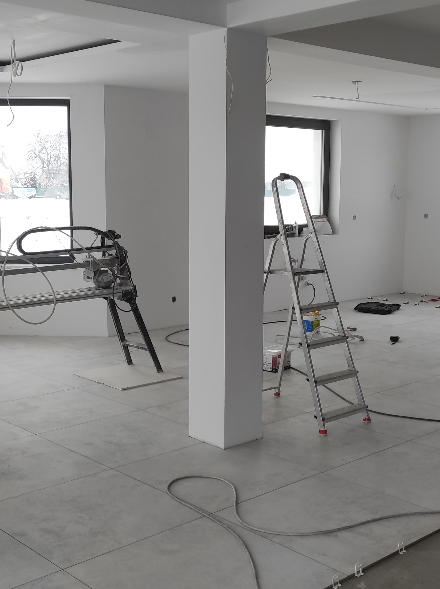 Wnętrze w trakcie wykańczania: szara podłoga z płytek, drabina aluminiowa, elektryczna przecinarka do płytek, widok przez okno na zimowy krajobraz.