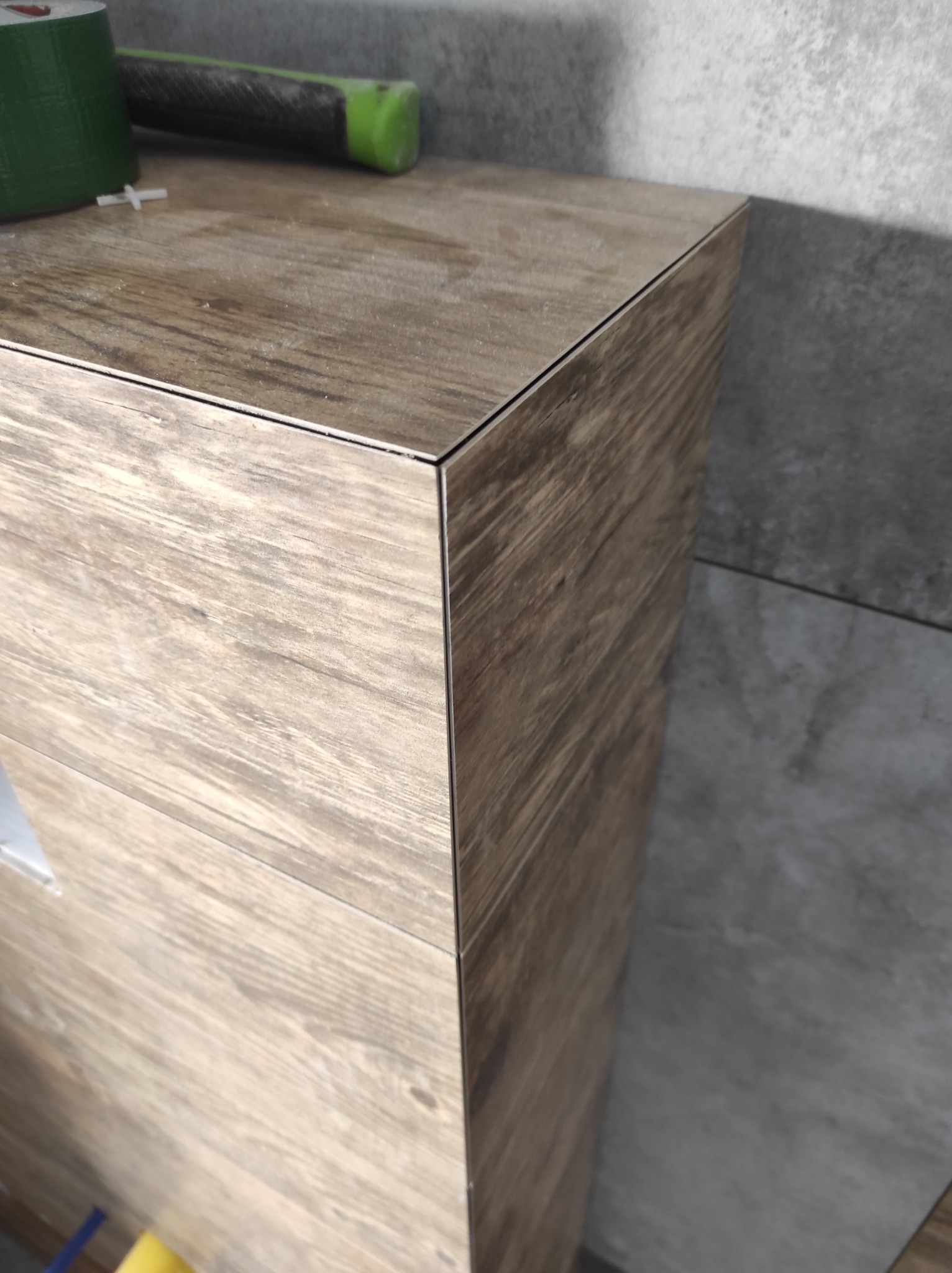 Wykończona obudowa WC w łazience z płytkami imitującymi drewno, widoczne narzędzia glazurnicze i fragment szarej ściany z płytek.