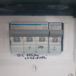 Tablica rozdzielcza z bezpiecznikami Hager, opisana odręcznie markerem, widoczne oznaczenia '1 ZAD', '2' oraz 'UPE GŁÓWNY ŁADOWARKI'.