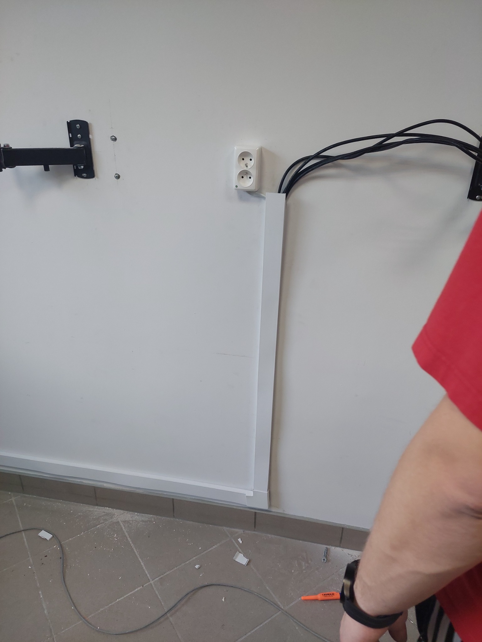 Biała ściana z zamontowanym uchwytem, gniazdkiem elektrycznym i wystającymi kablami, z widoczną listwą przypodłogową i fragmentem podłogi z resztkami po remoncie oraz fragmentem ręki w czerwonej...