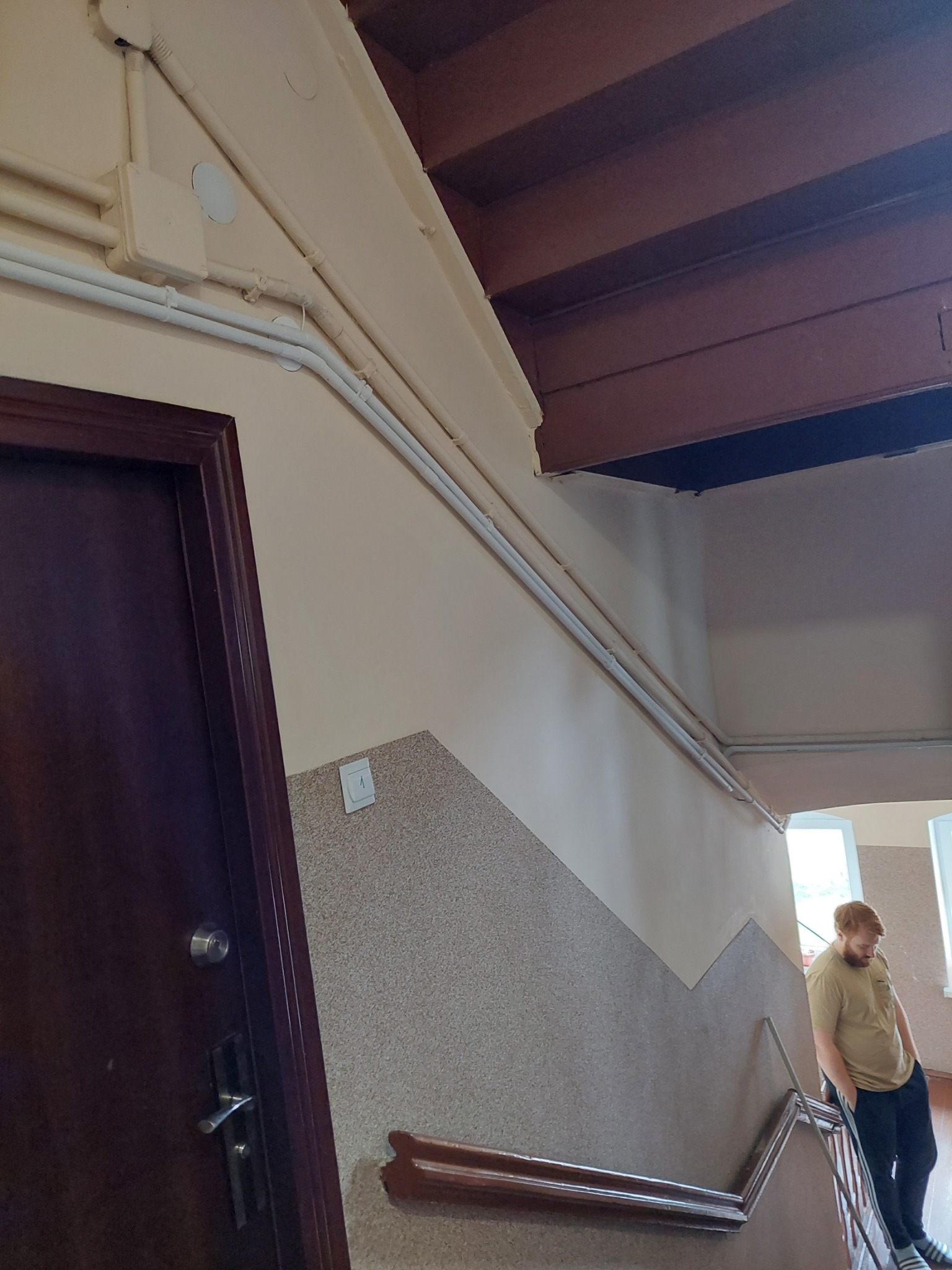Klatka schodowa z widocznymi rurami instalacji elektrycznej biegnącymi wzdłuż ściany, z osobą stojącą na schodach w tle.