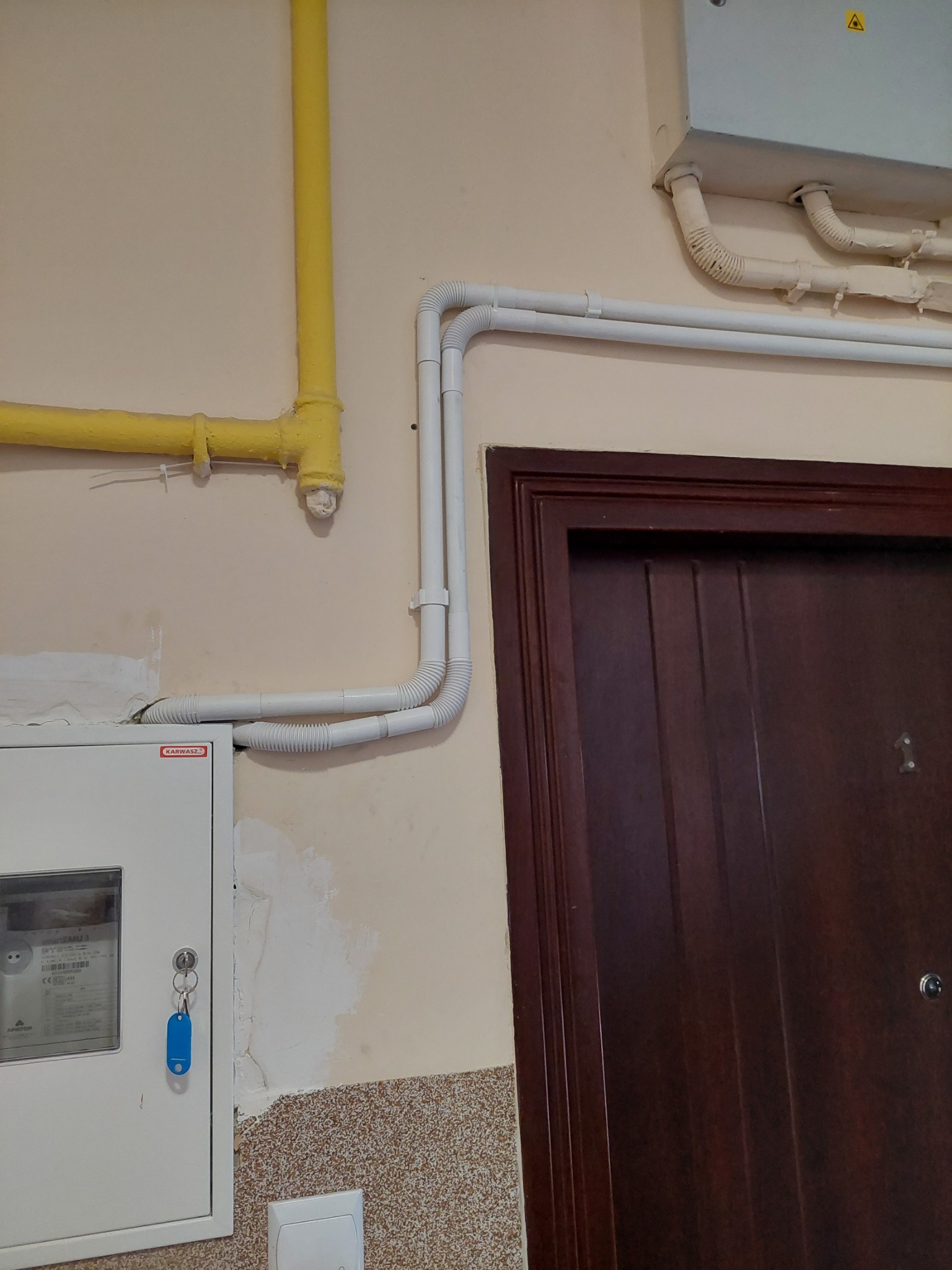 Żółta rura gazowa i białe rury osłonowe instalacji elektrycznej przy brązowych drzwiach wejściowych i białej skrzynce z licznikiem, widoczne ślady po naprawach na ścianie.