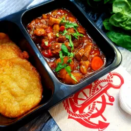 Danie w czarnym pojemniku: placki ziemniaczane i gulasz wołowy z warzywami, udekorowany pietruszką, obok sos śmietanowy i serwetka z motywem kotwicy.