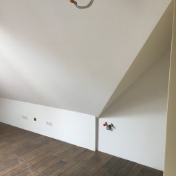Aleksander Tomaszczuk - Poddasze po remoncie: Świeżo pomalowane białe ściany skośne, drewniana podłoga, wystające gniazdka elektryczne i rury instalacyjne na ścianie.