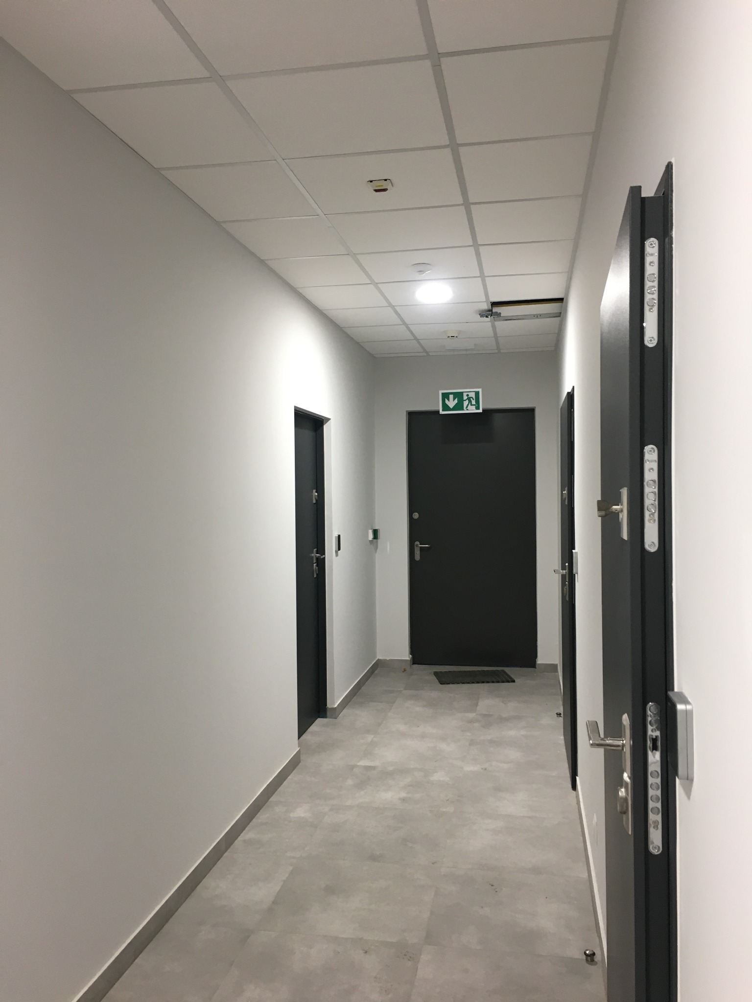 Korytarz biurowy z szarymi drzwiami i świeżo pomalowanymi białymi ścianami, widoczny znak ewakuacyjny.