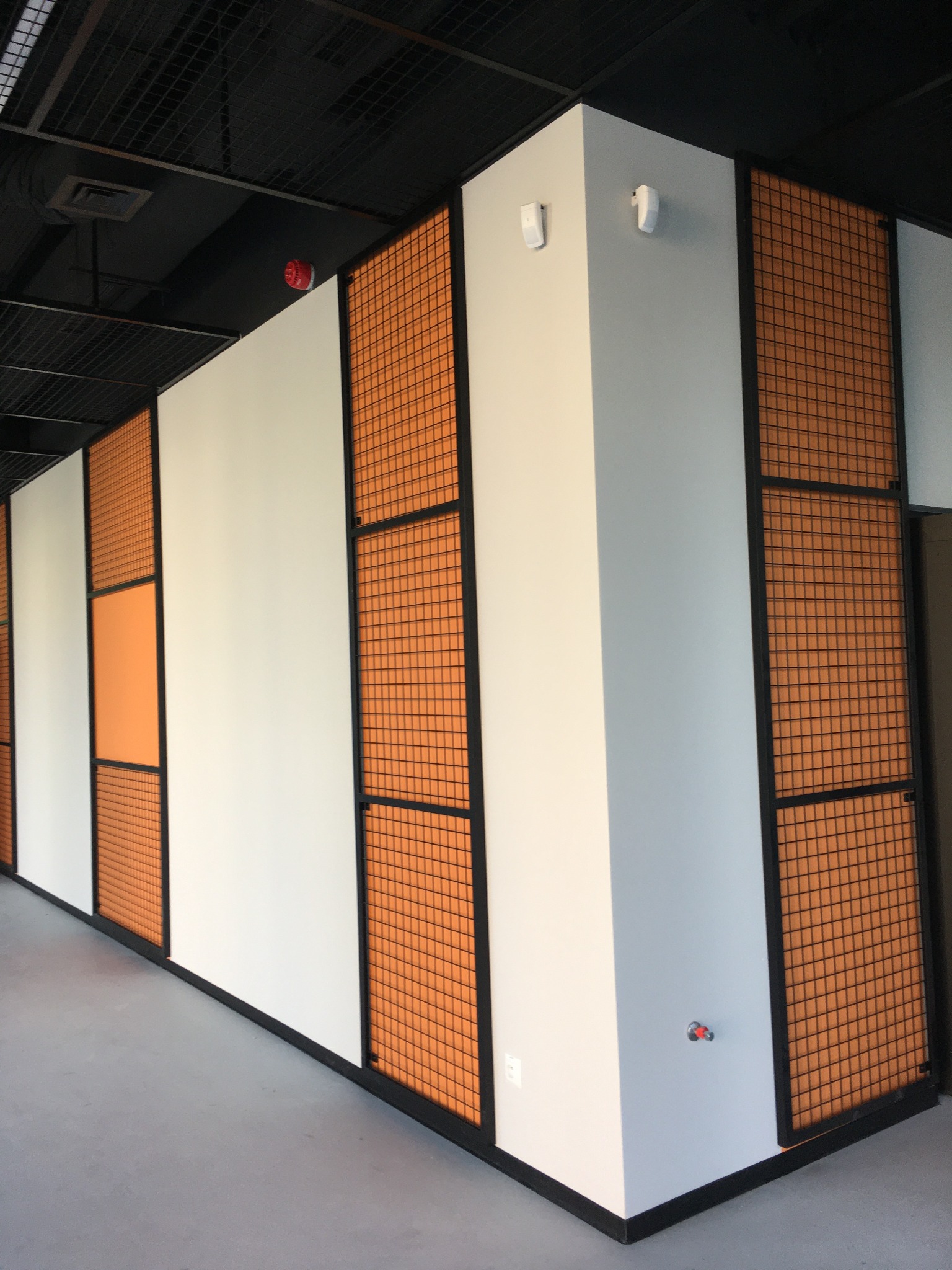 Białe i pomarańczowe panele ścienne z czarną metalową siatką w nowoczesnym biurze, widoczny fragment szarej podłogi i czarny sufit z kratką.