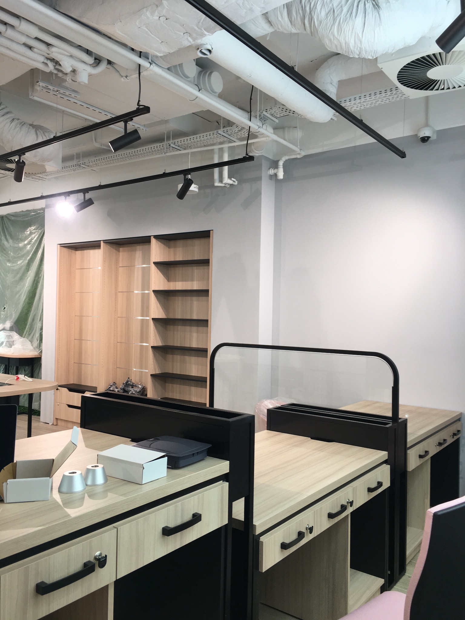 Nowoczesne biuro w trakcie przygotowania: biurka z jasnego drewna, czarne elementy konstrukcyjne, surowa biała ściana, widoczne instalacje pod sufitem, częściowo zasłonięte regały, rozpakowane...