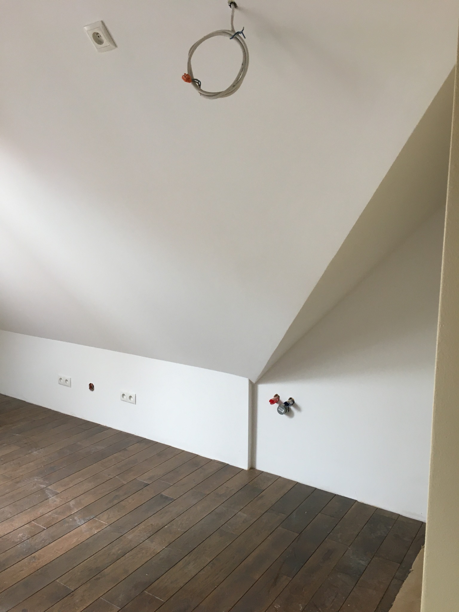 Poddasze po remoncie: Świeżo pomalowane białe ściany skośne, drewniana podłoga, wystające gniazdka elektryczne i rury instalacyjne na ścianie.