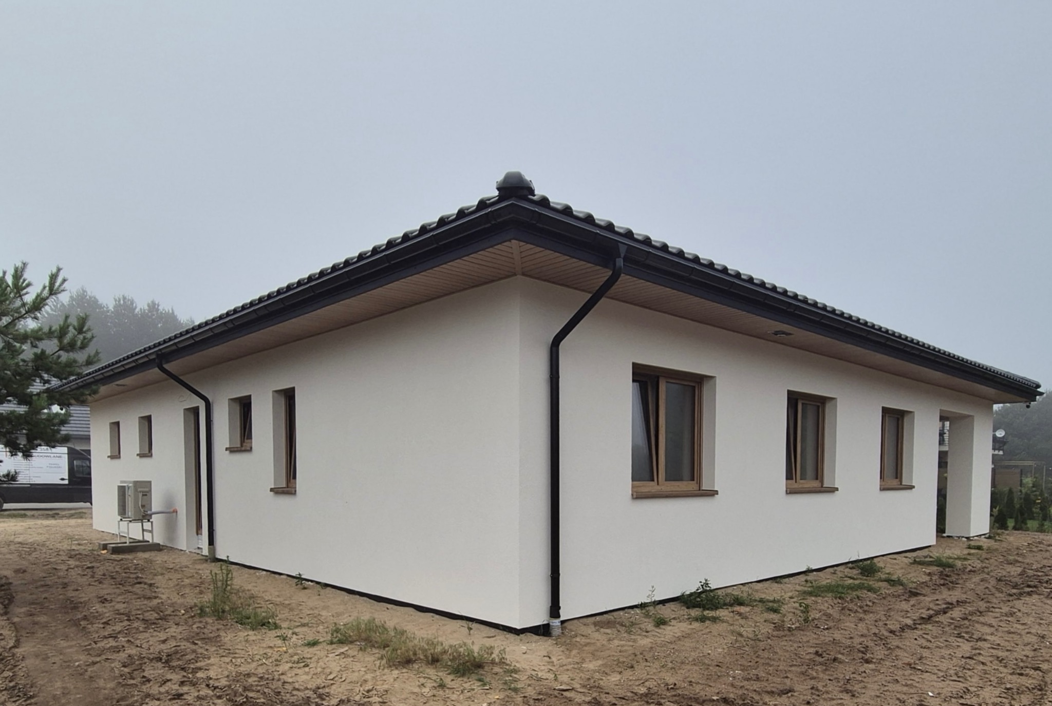 Parterowy dom z białą elewacją i ciemną dachówką w pochmurny dzień. Widoczne okna z drewnianymi ramami i czarne rynny. Minimalistyczny styl.