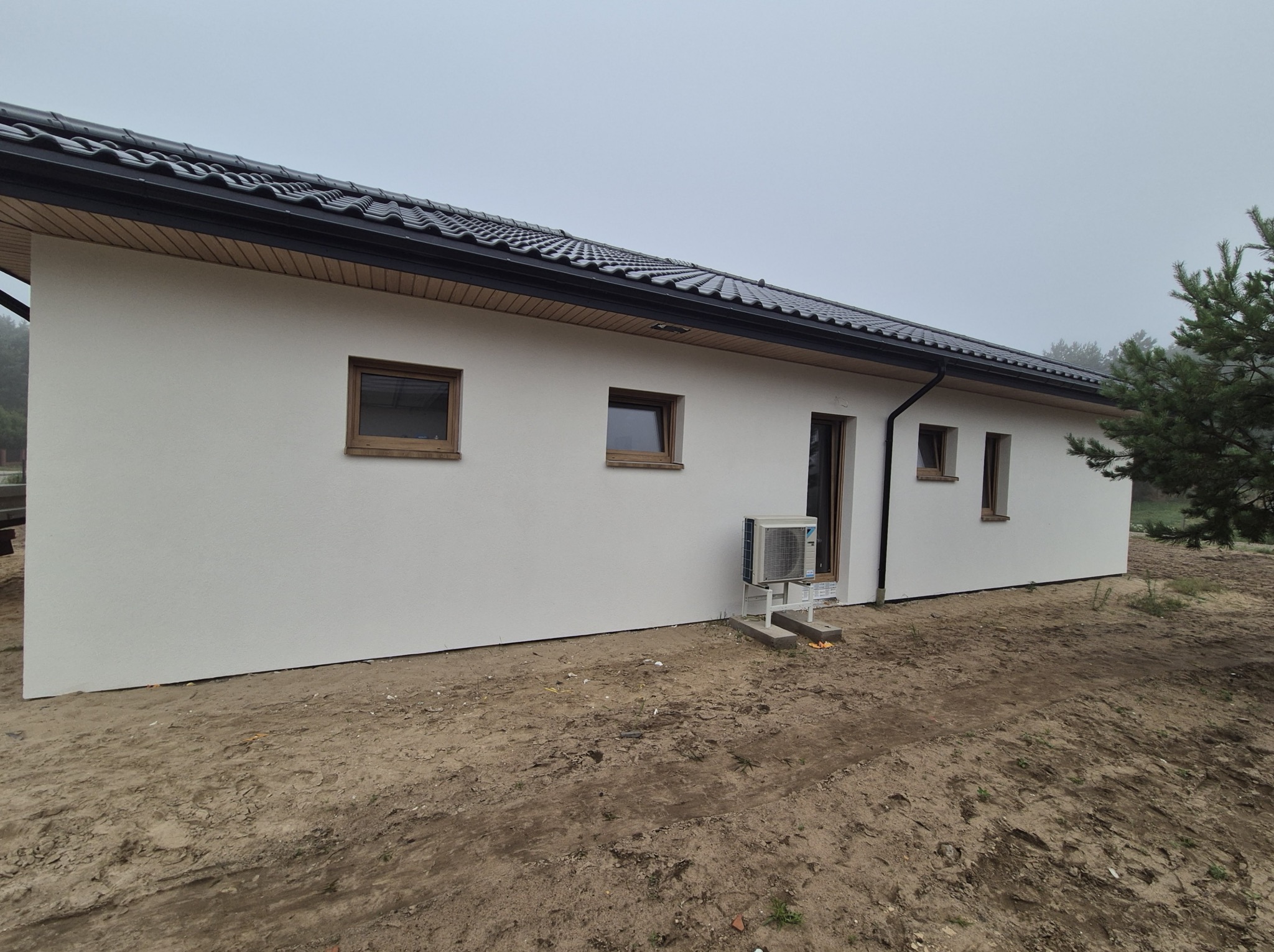 Boczny widok domu z białą elewacją, brązowymi oknami i ciemnym dachem. Widoczny klimatyzator na zewnątrz. Tło stanowi pochmurne niebo i fragment zieleni.