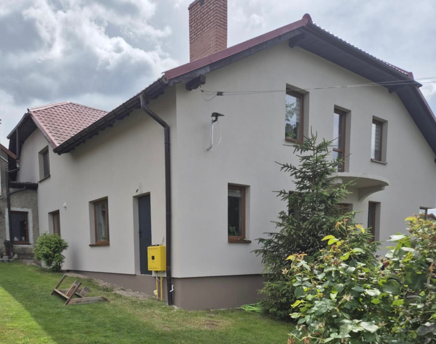 Dwukondygnacyjny dom z jasną elewacją, brązowymi ramami okiennymi i czerwonym dachem, widziany z perspektywy ogrodu z zielenią i żółtą skrzynką gazową.