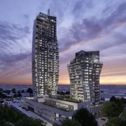 Dwa nowoczesne wieżowce o asymetrycznych bryłach, oświetlone wieczornym światłem, z widocznymi światłami w oknach, na tle zachodzącego słońca i zurbanizowanego krajobrazu.