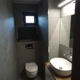 Nowoczesna łazienka w odcieniach szarości z wiszącą toaletą, grzejnikiem żeberkowym pod oknem i umywalką nablatową na drewnianym blacie.