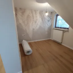 Poddasze po remoncie z tapetą w liście, drewnianą podłogą i zwiniętym dywanem w rogu, widok na okno z grzejnikiem.