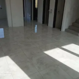 Jasne, błyszczące płytki podłogowe w przestronnym wnętrzu w stanie deweloperskim, z widocznymi promieniami słońca padającymi przez okno, częściowo wykończone schody i widoczne niedoskonałości...