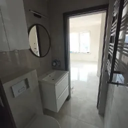 Wykończona łazienka z szarą glazurą, wiszącą toaletą, szafką z umywalką i okrągłym lustrem, widok na jasne pomieszczenie przez otwarte drzwi.