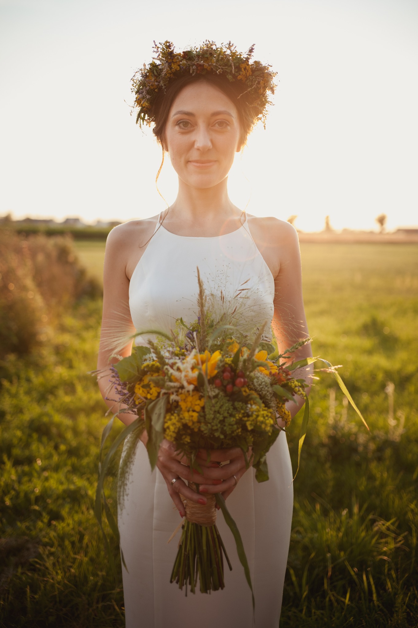 Panna młoda w białej sukni i wianku z polnych kwiatów trzyma bukiet w rustykalnym stylu na tle łąki o zachodzie słońca.