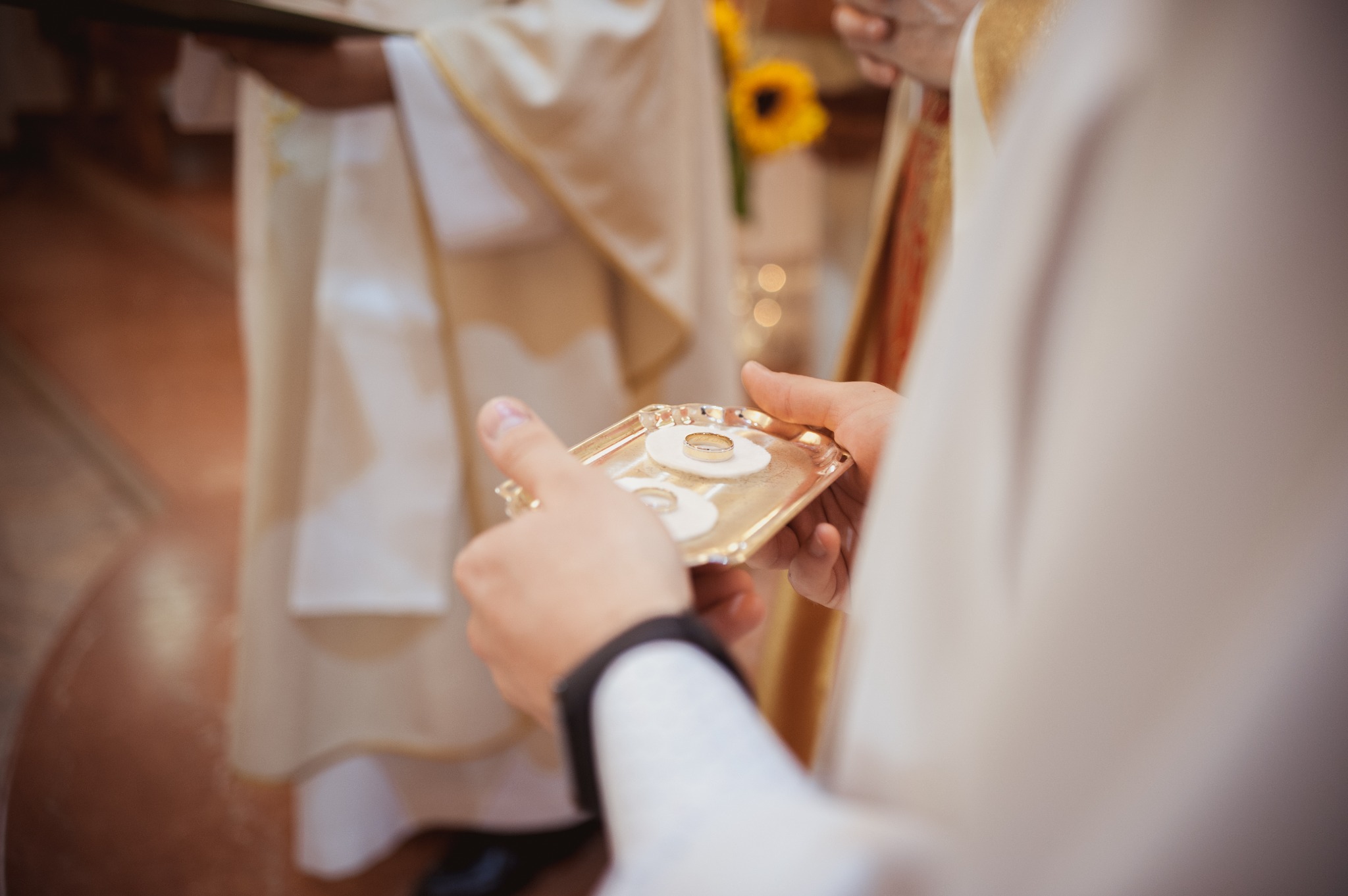 Złote obrączki na złotej tacy z hostiami, trzymane przez osobę w białej szacie podczas ceremonii ślubnej w kościele, z widocznym fragmentem żółtego słonecznika w tle.