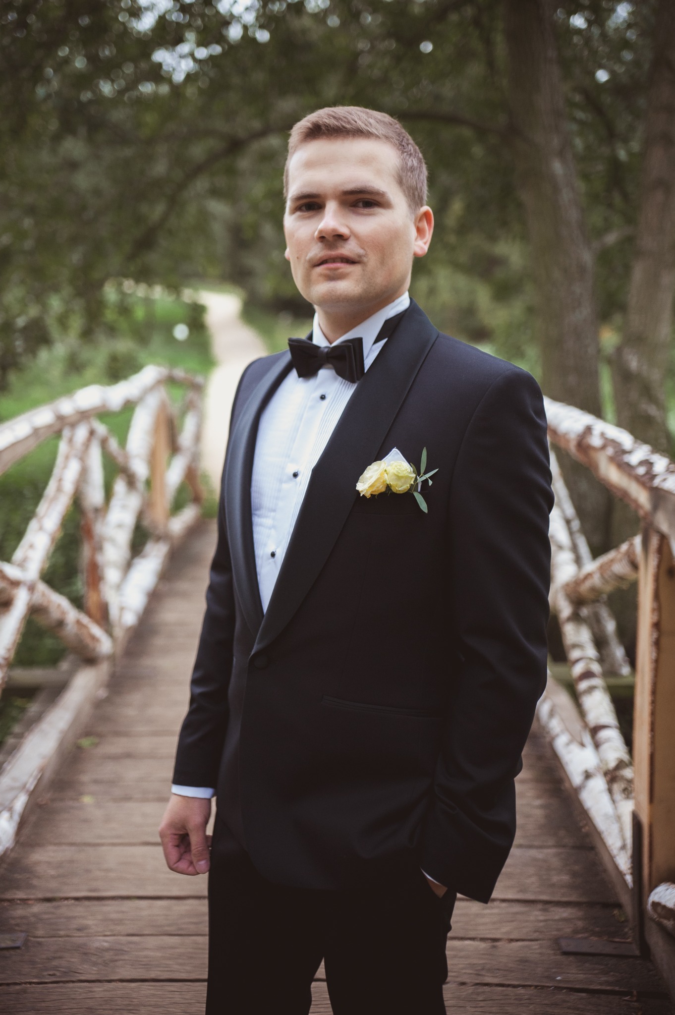Mężczyzna w czarnym smokingu z żółtą butonierką na drewnianym mostku w zalesionym otoczeniu.