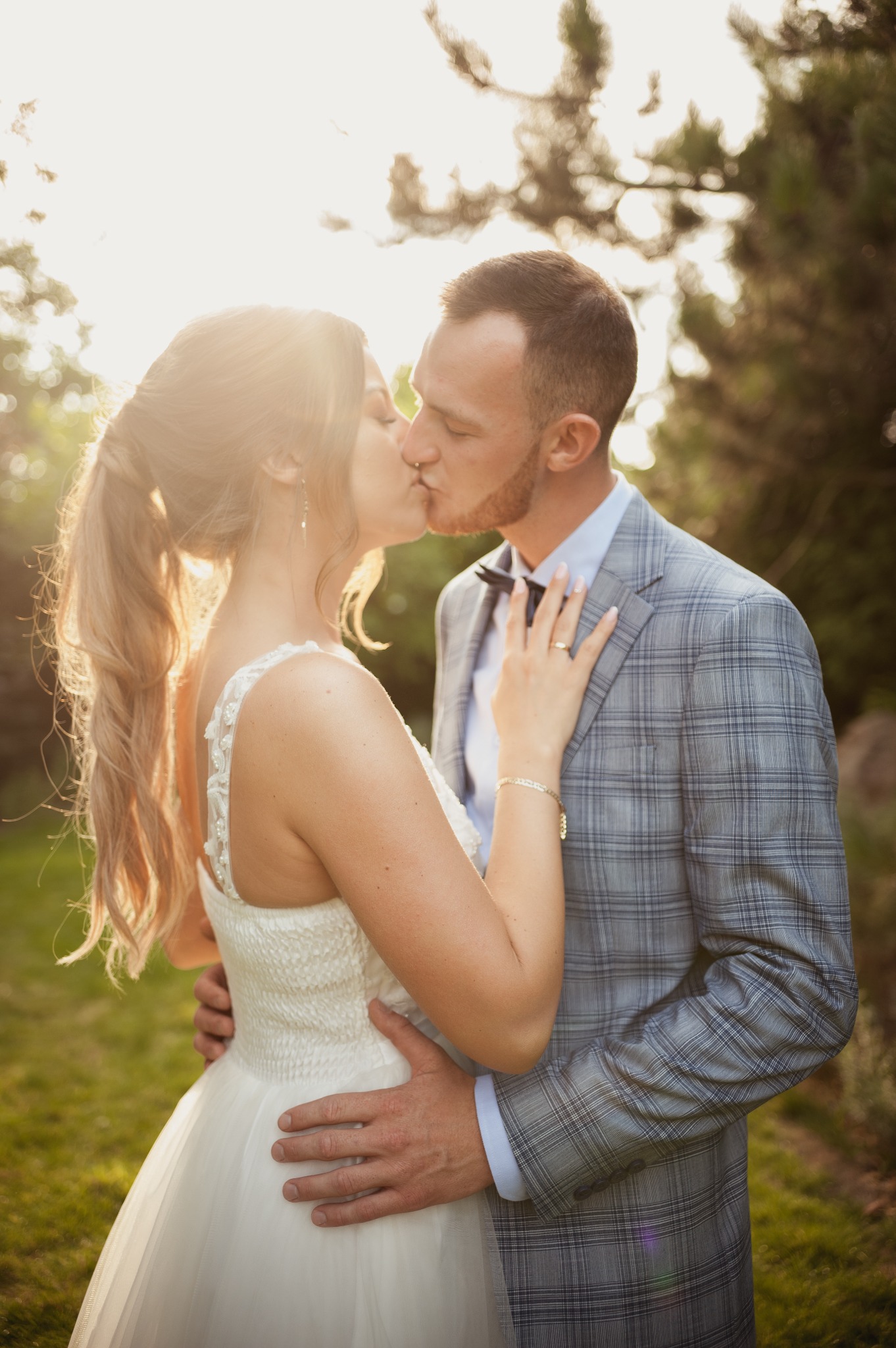 Pocałunek pary młodej w plenerze, panna młoda w sukni z koronkowymi ramiączkami i upiętymi włosami, pan młody w szarej kraciastej marynarce i muszce, ciepłe światło słoneczne.