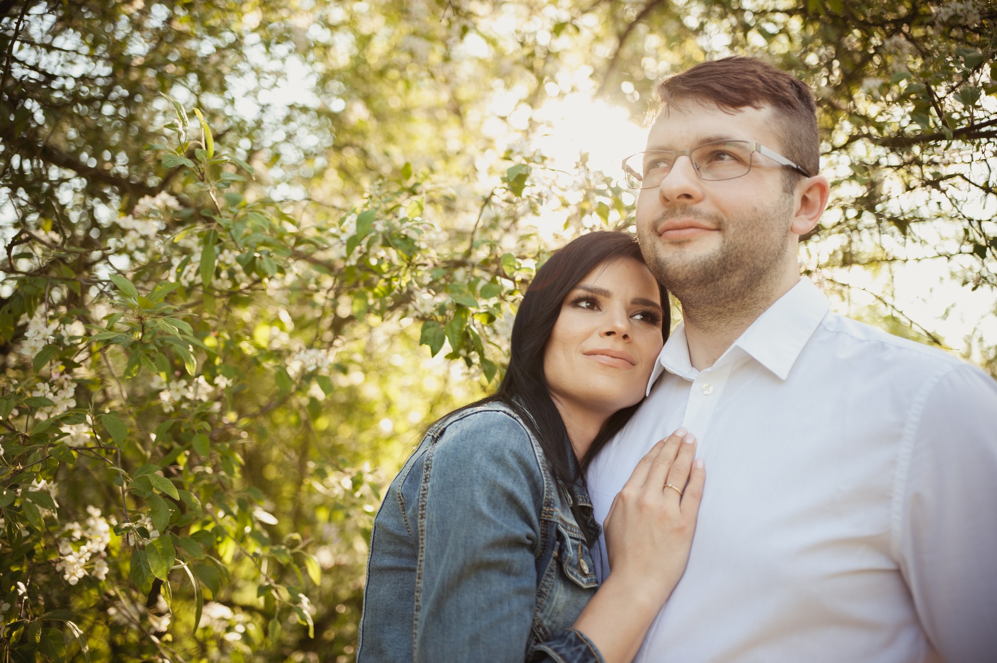 Zakochana para przytulona na tle kwitnących drzew, kobieta w dżinsowej kurtce z pierścionkiem na palcu, mężczyzna w okularach i białej koszuli, delikatne światło słoneczne.