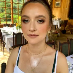 Portret kobiety z profesjonalnym makijażem: brązowe cienie, podkreślone rzęsy, delikatny róż na policzkach i matowe usta w odcieniu nude, na tle drewnianego wnętrza z zastawionym stołem.