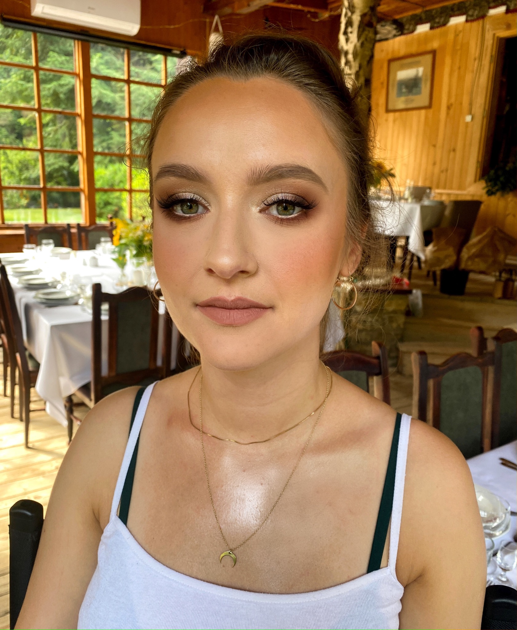 Portret kobiety z profesjonalnym makijażem: brązowe cienie, podkreślone rzęsy, delikatny róż na policzkach i matowe usta w odcieniu nude, na tle drewnianego wnętrza z zastawionym stołem.