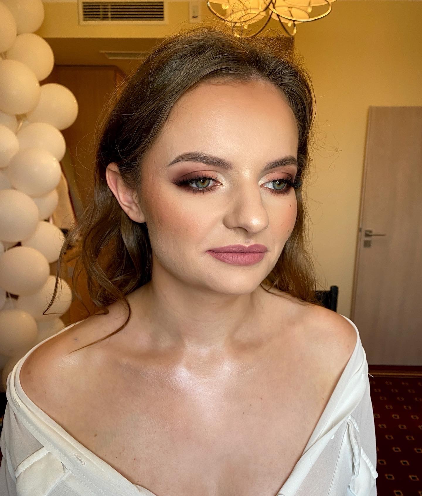 Portret kobiety z profesjonalnie wykonaną stylizacją oczu w odcieniach brązu i różu, oraz matowymi ustami w kolorze nude. W tle widoczne białe balony i fragment pomieszczenia.