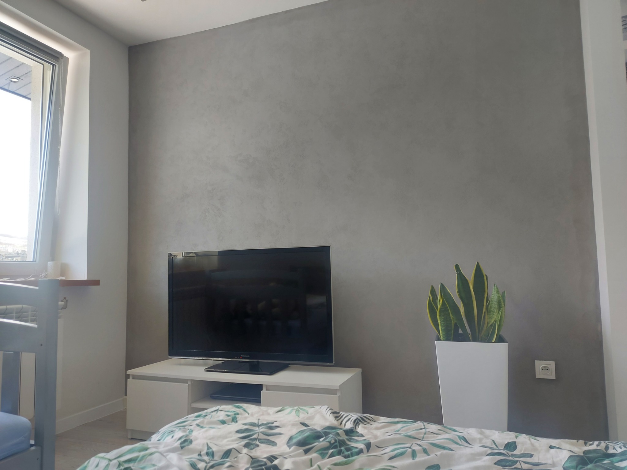 Pokój z szarą ścianą o nieregularnej fakturze, telewizorem na białej szafce RTV, rośliną w białej donicy i łóżkiem w tropikalne wzory.