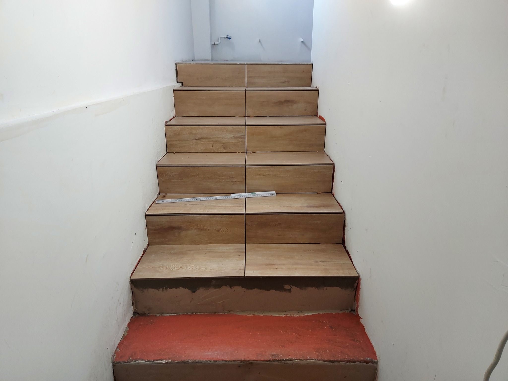 Schody wyłożone panelami drewnopodobnymi w trakcie montażu, widoczne niedociągnięcia przy ścianach i czerwona farba na dolnym stopniu, na jednym ze stopni leży miarka.