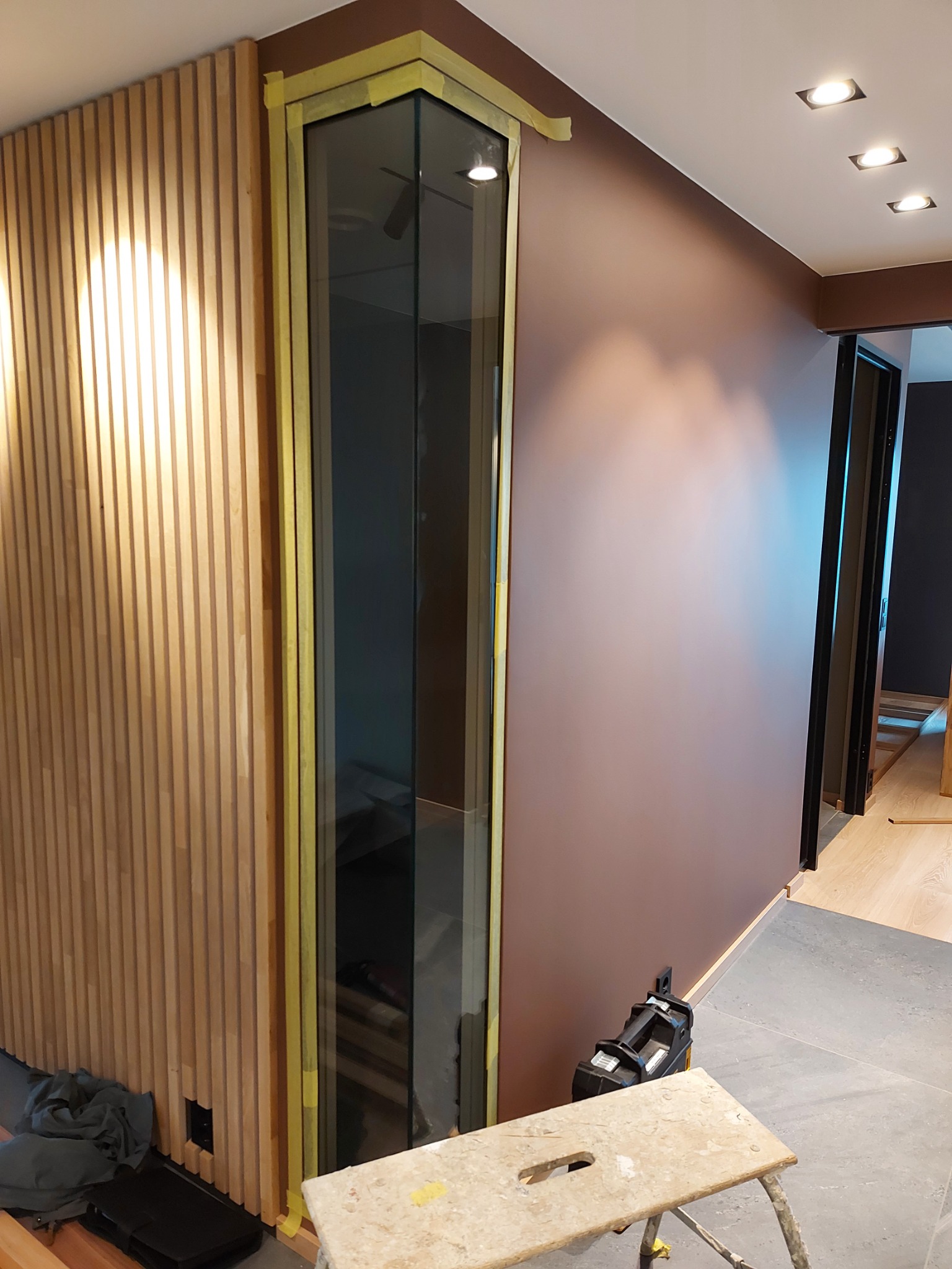 Wnętrze w trakcie prac wykończeniowych: drewniana ściana z lamelami, narożne okno zabezpieczone taśmą malarską, pomalowana ściana w kolorze brązu, widoczny stołek malarski i lampa budowlana.