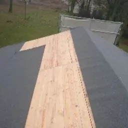 Fragment dachu pokryty papą, z widocznym, niezabezpieczonym fragmentem drewnianej konstrukcji, ukazującym etap prac dekarskich, z zielonym terenem w tle.