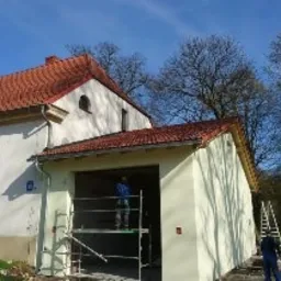 Prace na wysokościach przy elewacji budynku: robotnik na rusztowaniu maluje ścianę na jasnożółty kolor, obok drabina, budynek z czerwonym dachem, w tle drzewa i błękitne niebo.