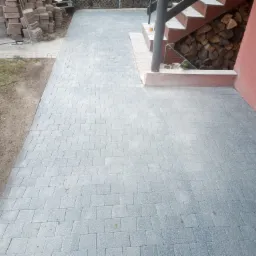 Świeżo ułożona, szara kostka brukowa prowadząca do schodów z drewnem opałowym pod nimi, widoczna ubijarka wibracyjna i stos kostki obok.