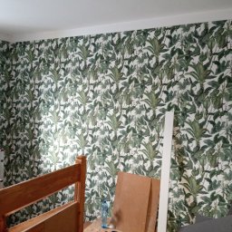 Firma Ogólnobudowlana Arkos - Ściana pokryta tapetą w motywy roślinne, widoczne fragmenty drewnianej ramy łóżka, butelka wody i elementy wykończeniowe, sugerujące prace remontowe w pokoju.