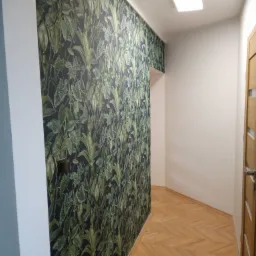 Wnętrze korytarza z tapetą w motywy roślinne, parkietem w jodełkę i drewnianymi drzwiami z przeszkleniem, widok z perspektywy podłogi.