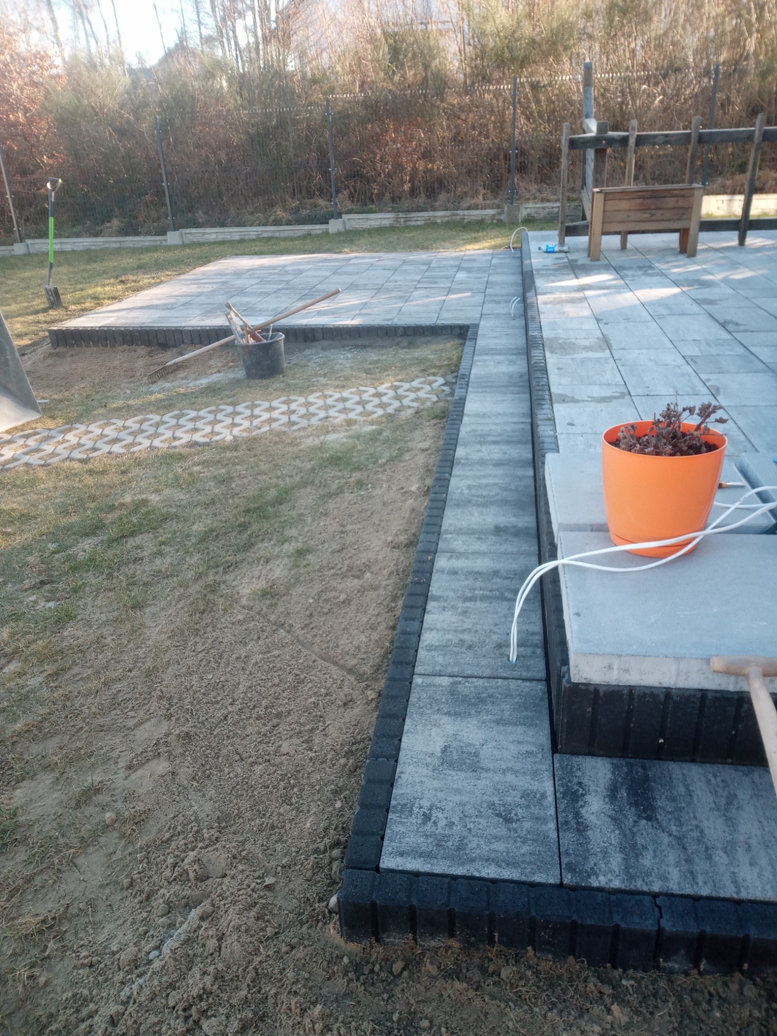 Fragment tarasu z jasnoszarej kostki brukowej obramowanej ciemnoszarą kostką, na skraju widoczna ziemia i trawa, w tle płot i drzewa.