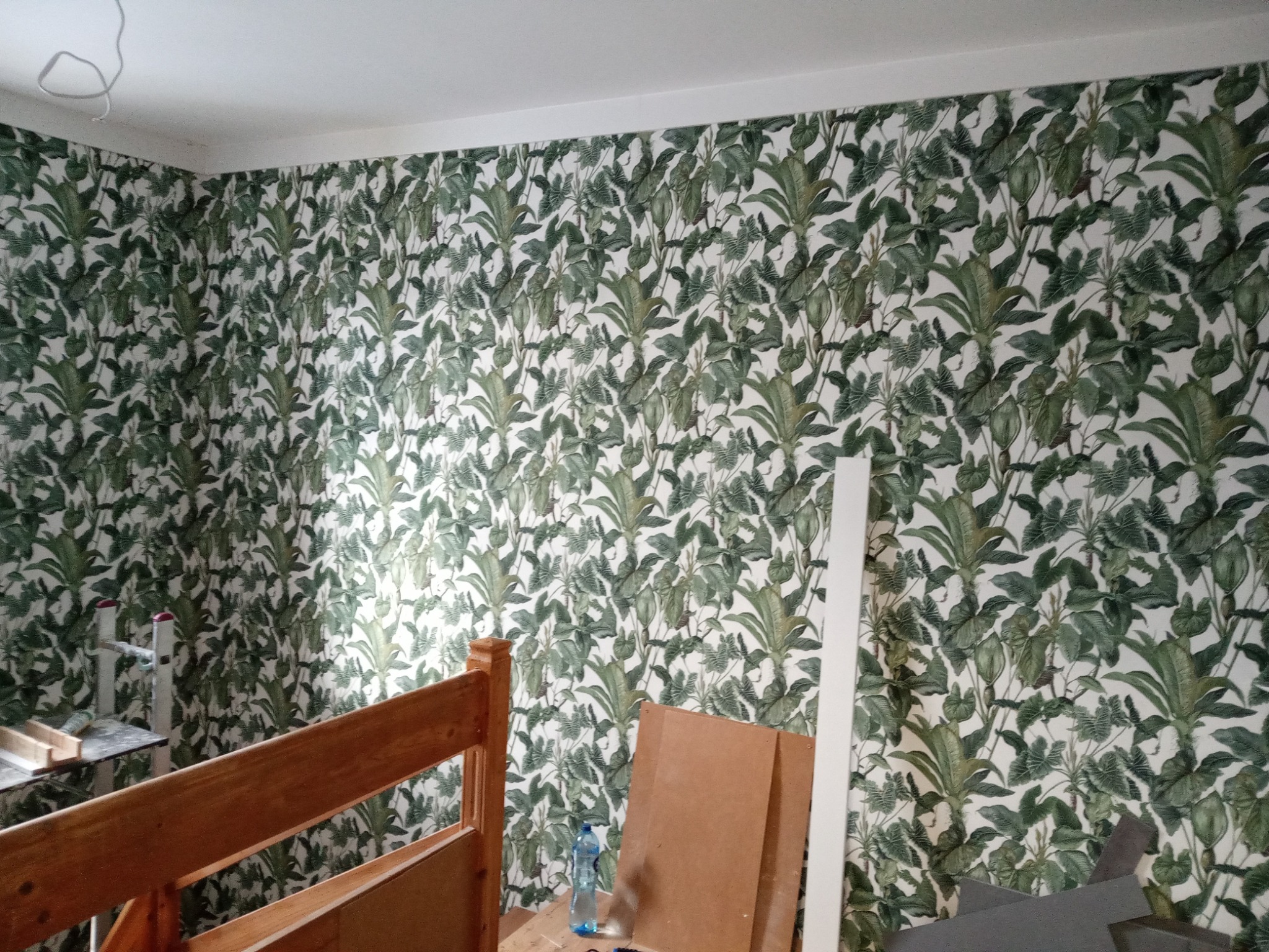 Ściana pokryta tapetą w motywy roślinne, widoczne fragmenty drewnianej ramy łóżka, butelka wody i elementy wykończeniowe, sugerujące prace remontowe w pokoju.
