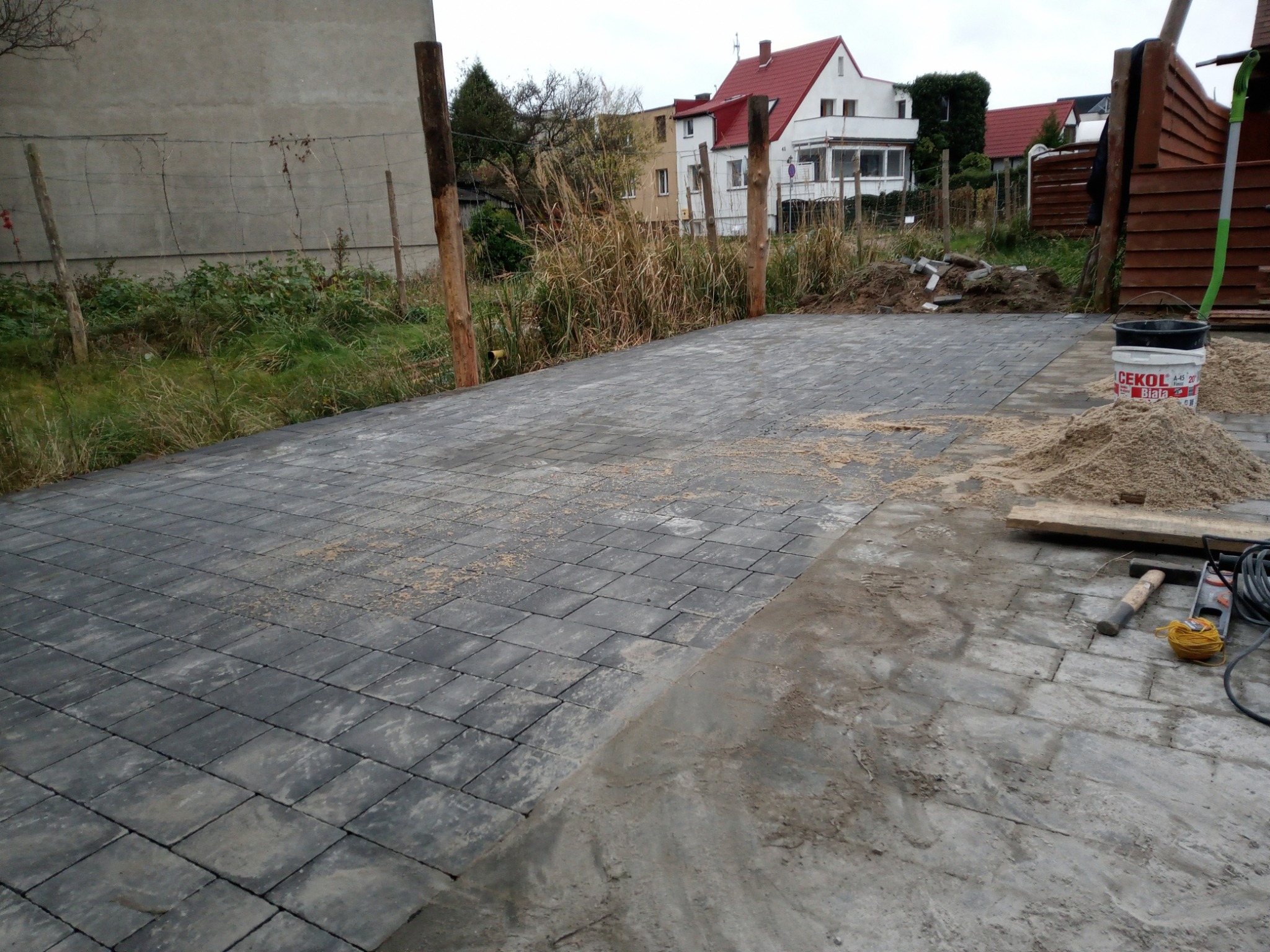 Ułożona szara kostka brukowa na posesji, widoczny fragment terenu przygotowany pod dalsze prace, narzędzia i wiadro z zaprawą, w tle budynki mieszkalne.