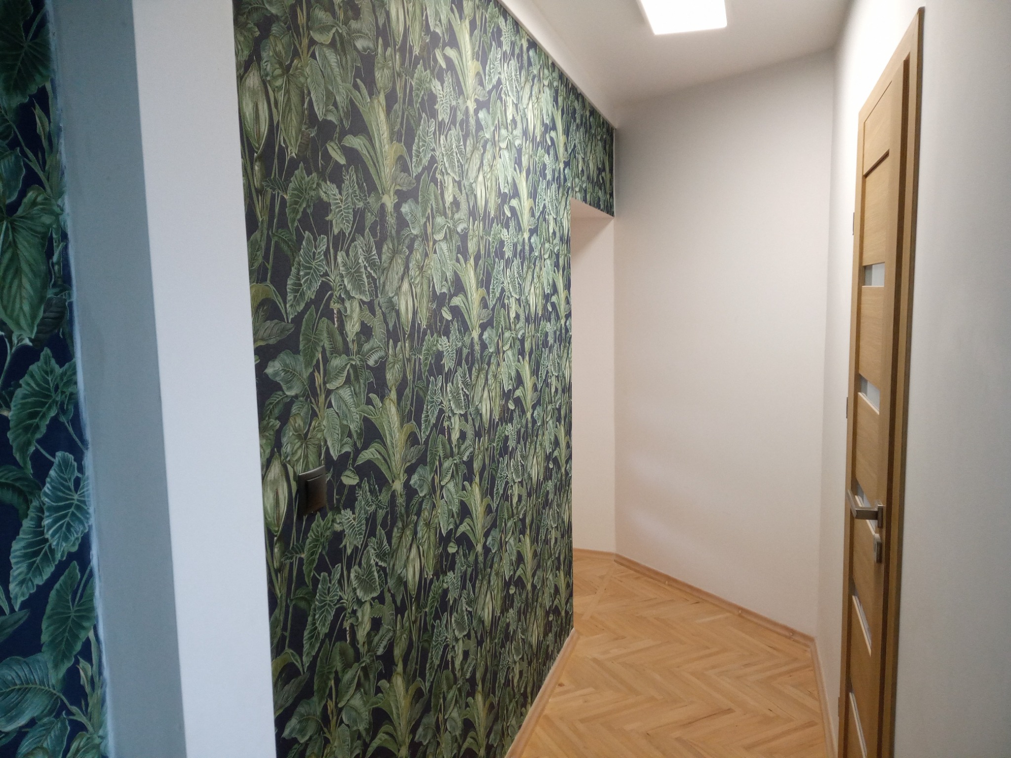 Wnętrze korytarza z tapetą w motywy roślinne, parkietem w jodełkę i drewnianymi drzwiami z przeszkleniem, widok z perspektywy podłogi.