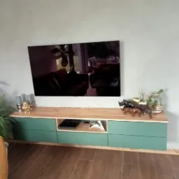 Nowoczesna szafka RTV z zielonymi frontami i drewnianym blatem, udekorowana figurkami zwierząt i świecznikami, z telewizorem na ścianie. Widoczny fragment sofy w kolorze musztardowym.