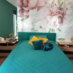 Sypialnia z turkusowym łóżkiem, drewnianymi szafkami nocnymi z czarnymi akcentami i fototapetą w kwiaty piwonii na ścianie.